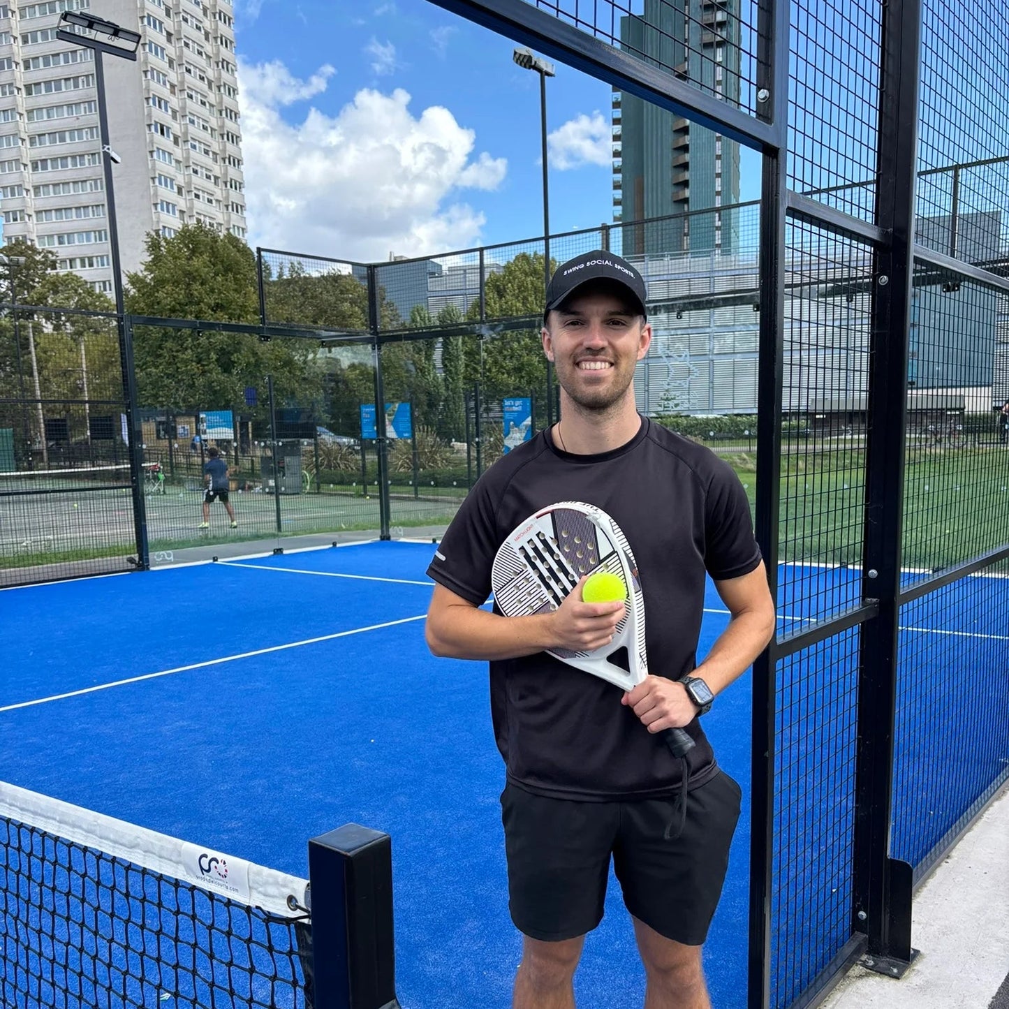 Man holding a paddle and ball on a blue padel court with buildings in the background